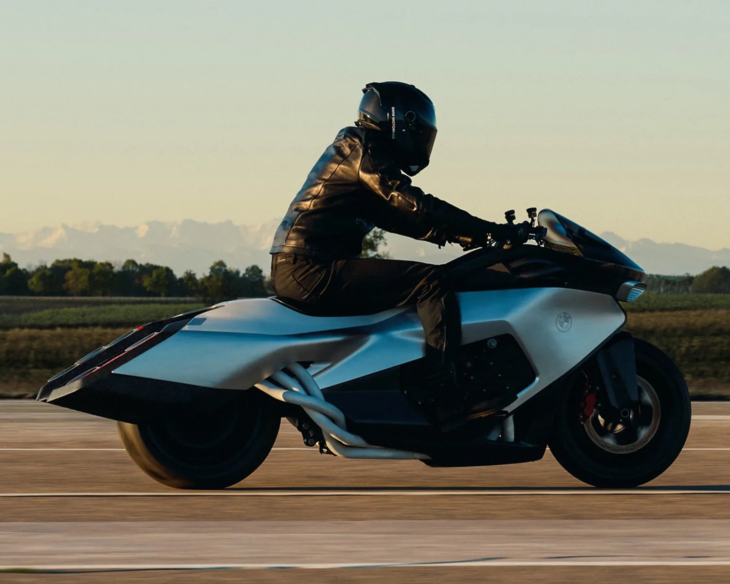 Person in black helmet and leather jacket riding a futuristic silver and black BMW motorcycle on a road.