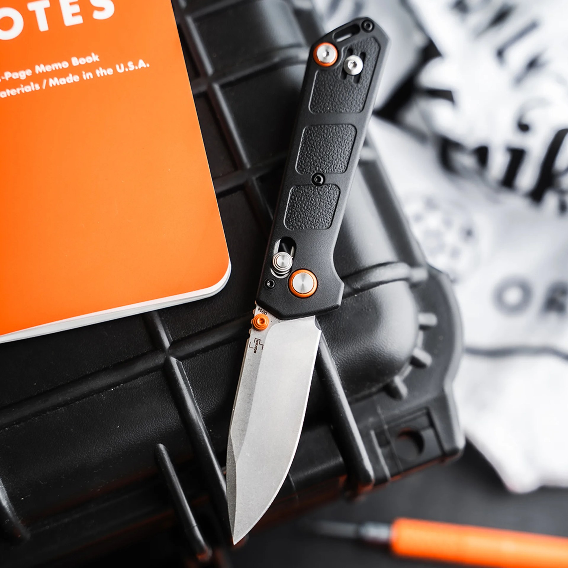 Folding knife with black textured handle and silver blade on a black surface next to an orange notebook.