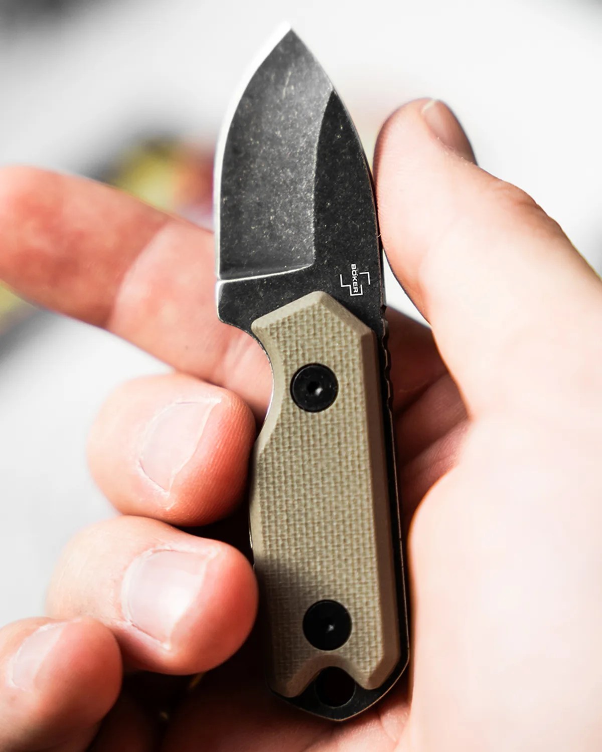 Hand holding a small Boker folding knife with a black blade and tan textured handle.