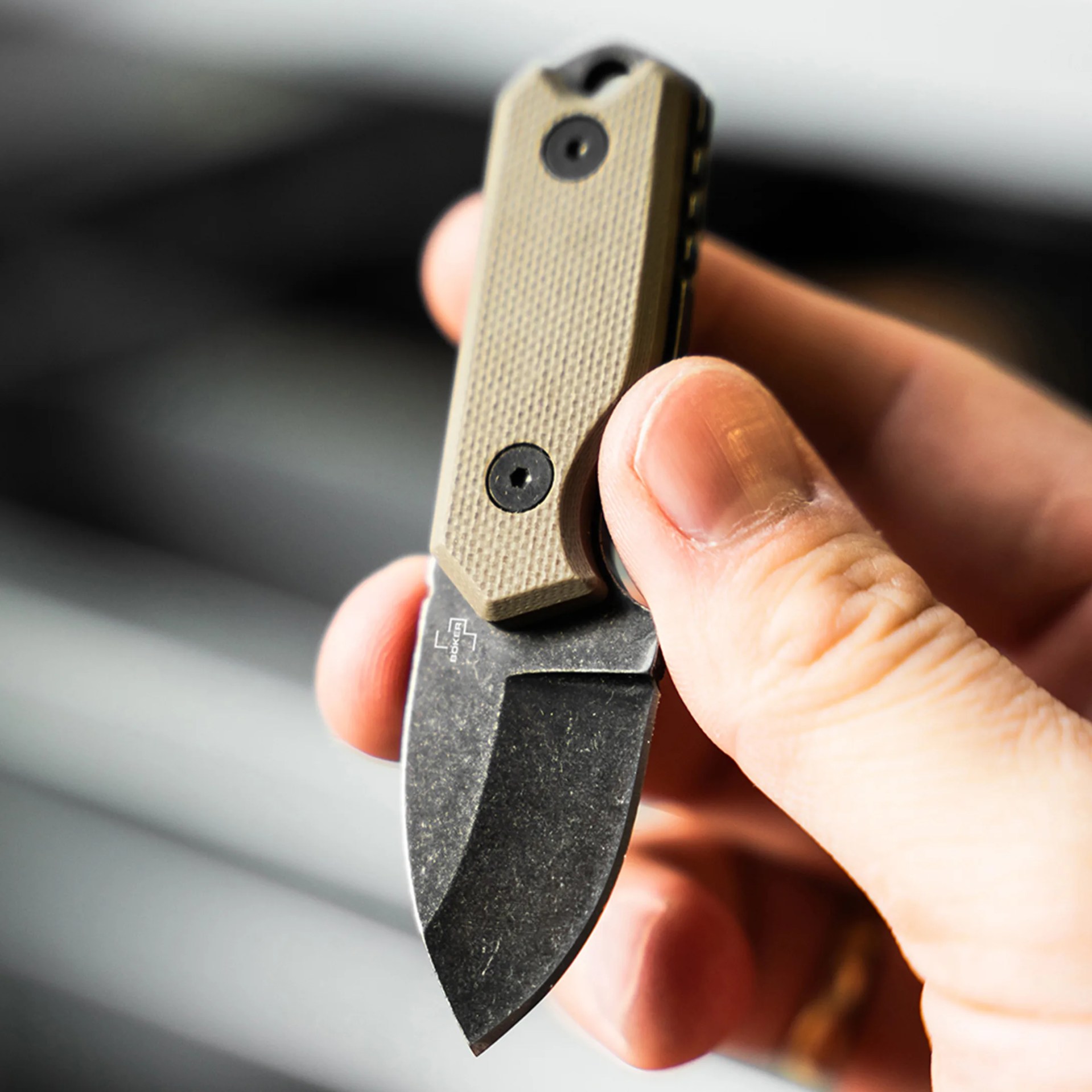 Hand holding a small folding knife with a textured tan handle and a black stonewashed blade.