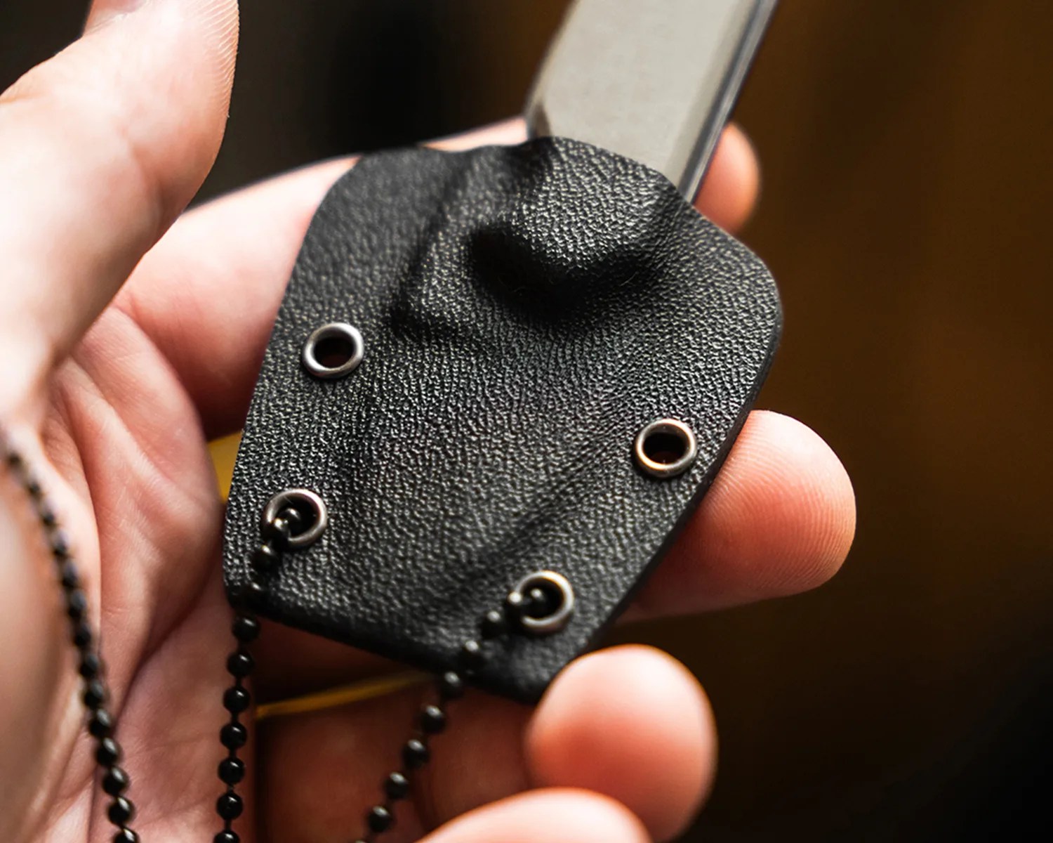 Hand holding a black textured sheath with metal eyelets and a chain attached.