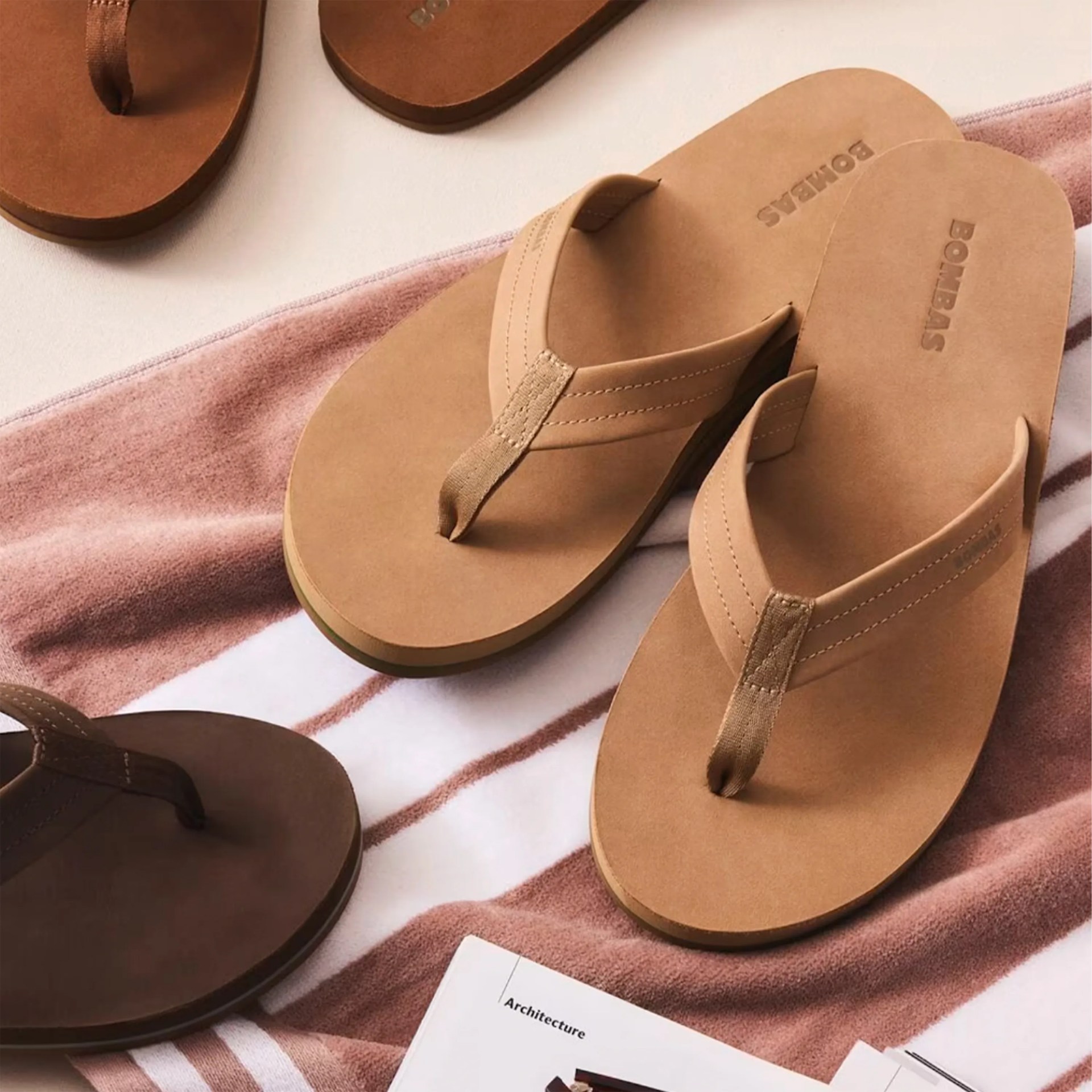 Pair of tan leather Bombas flip-flops on a striped towel next to a magazine.