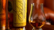 Close-up of a whiskey bottle with a yellow label and a whiskey glass partially filled with amber liquid.