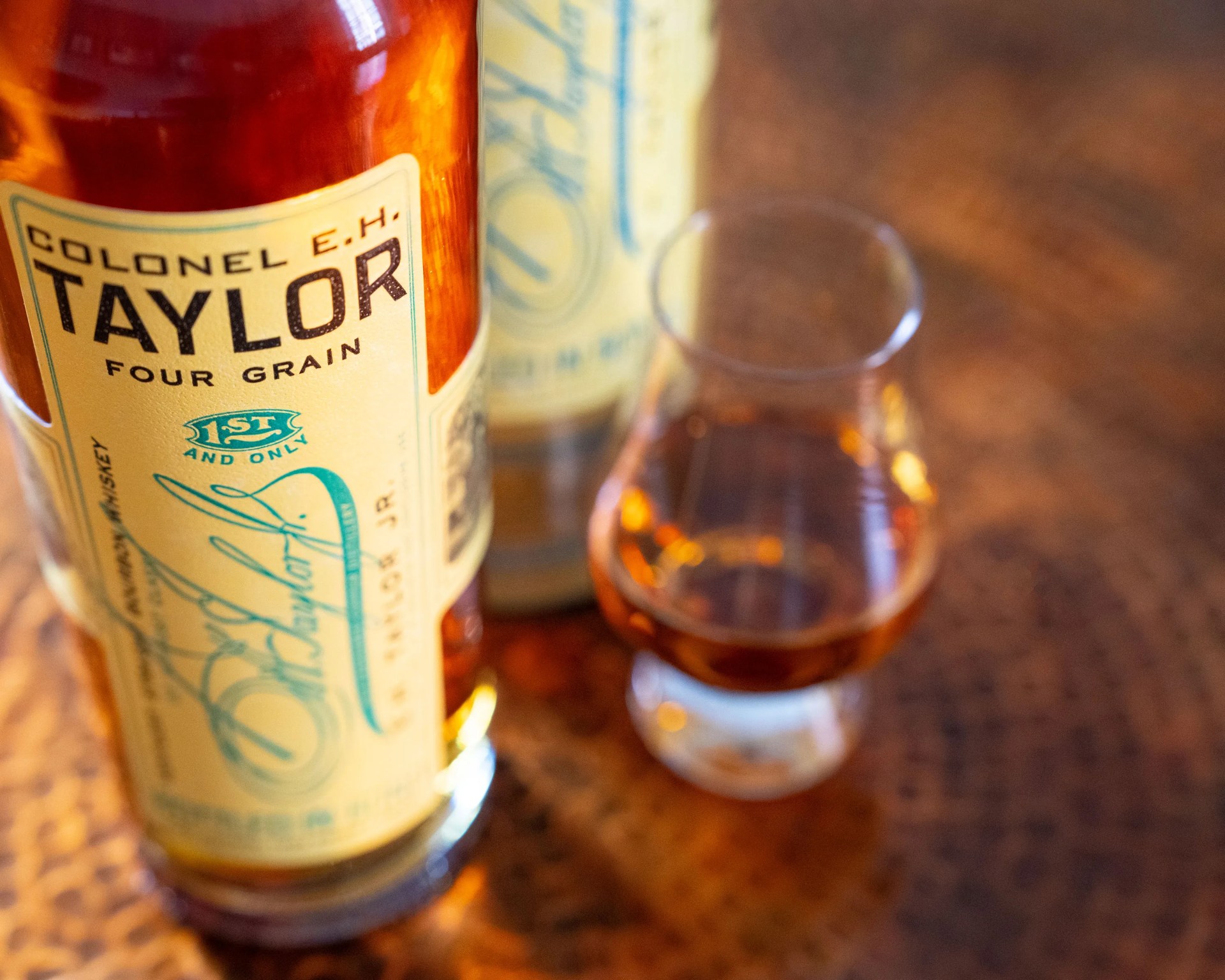 Amber whiskey bottle labeled "Colonel E.H. Taylor Four Grain" next to a whiskey glass on a wooden surface.