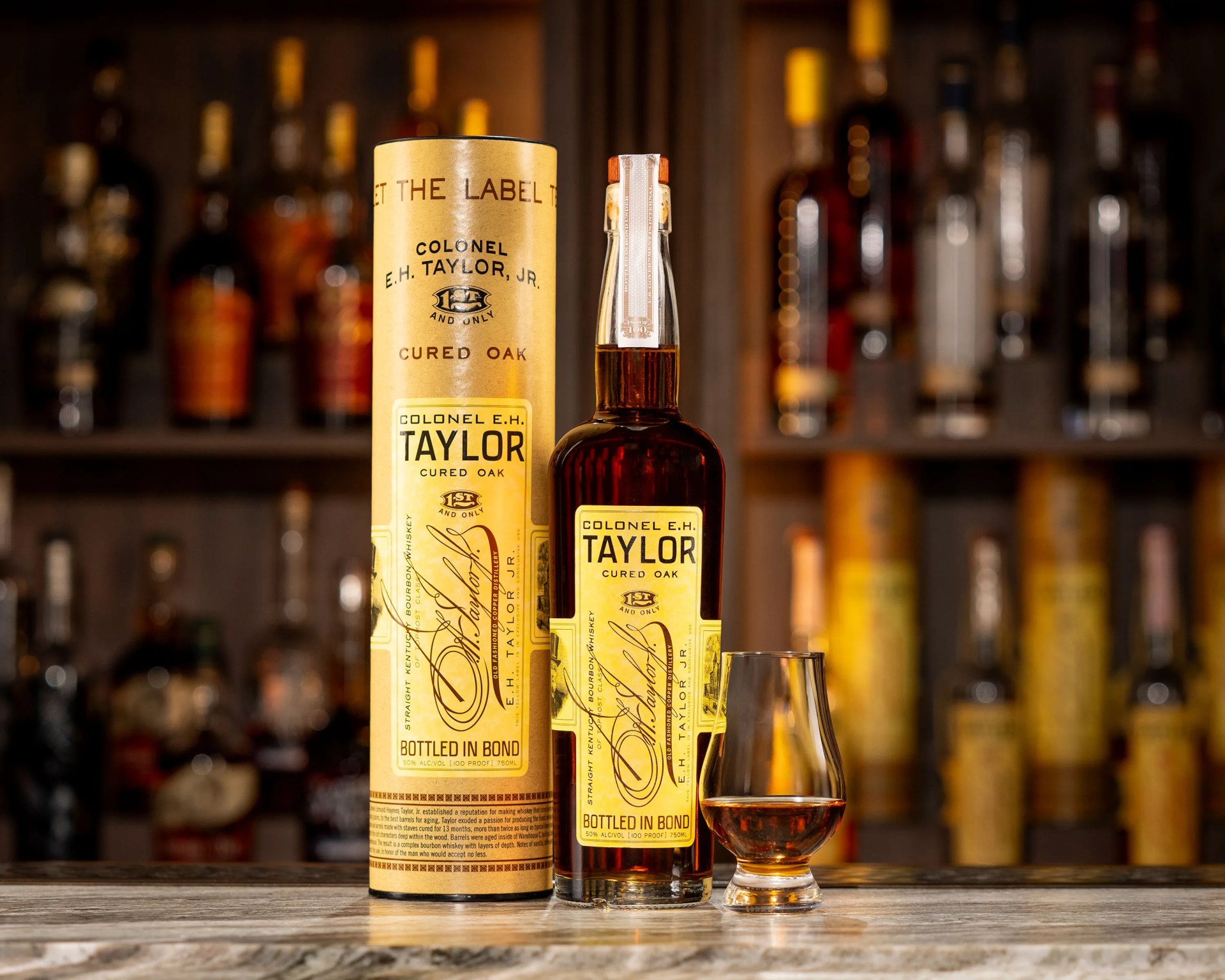 Bottle and cylindrical container of Colonel E.H. Taylor Jr. Cured Oak bourbon whiskey with a filled whiskey glass on a wooden surface.