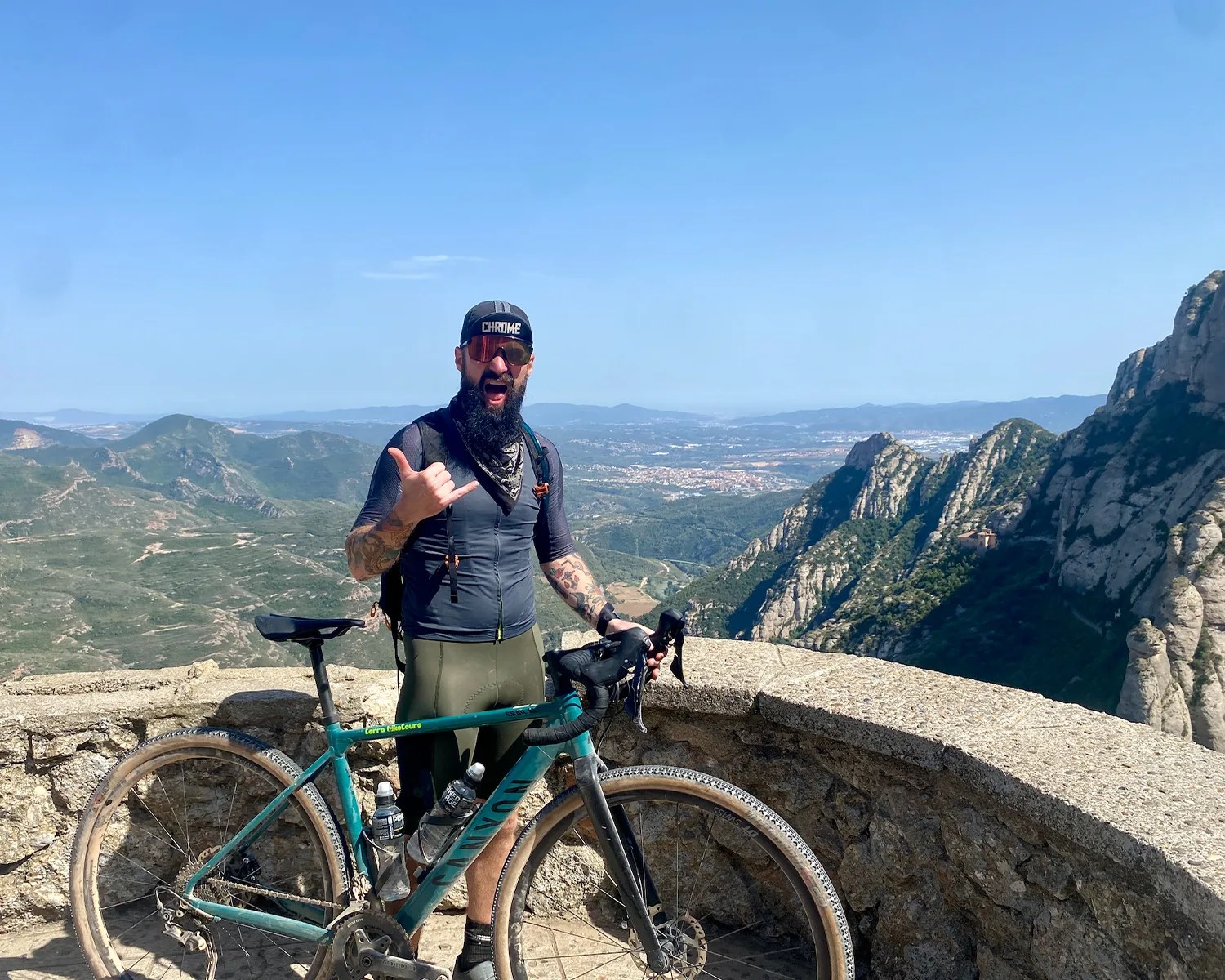 Man with a beard and tattoos wearing cycling gear and sunglasses standing with a teal Canyon bike on a mountain overlook.