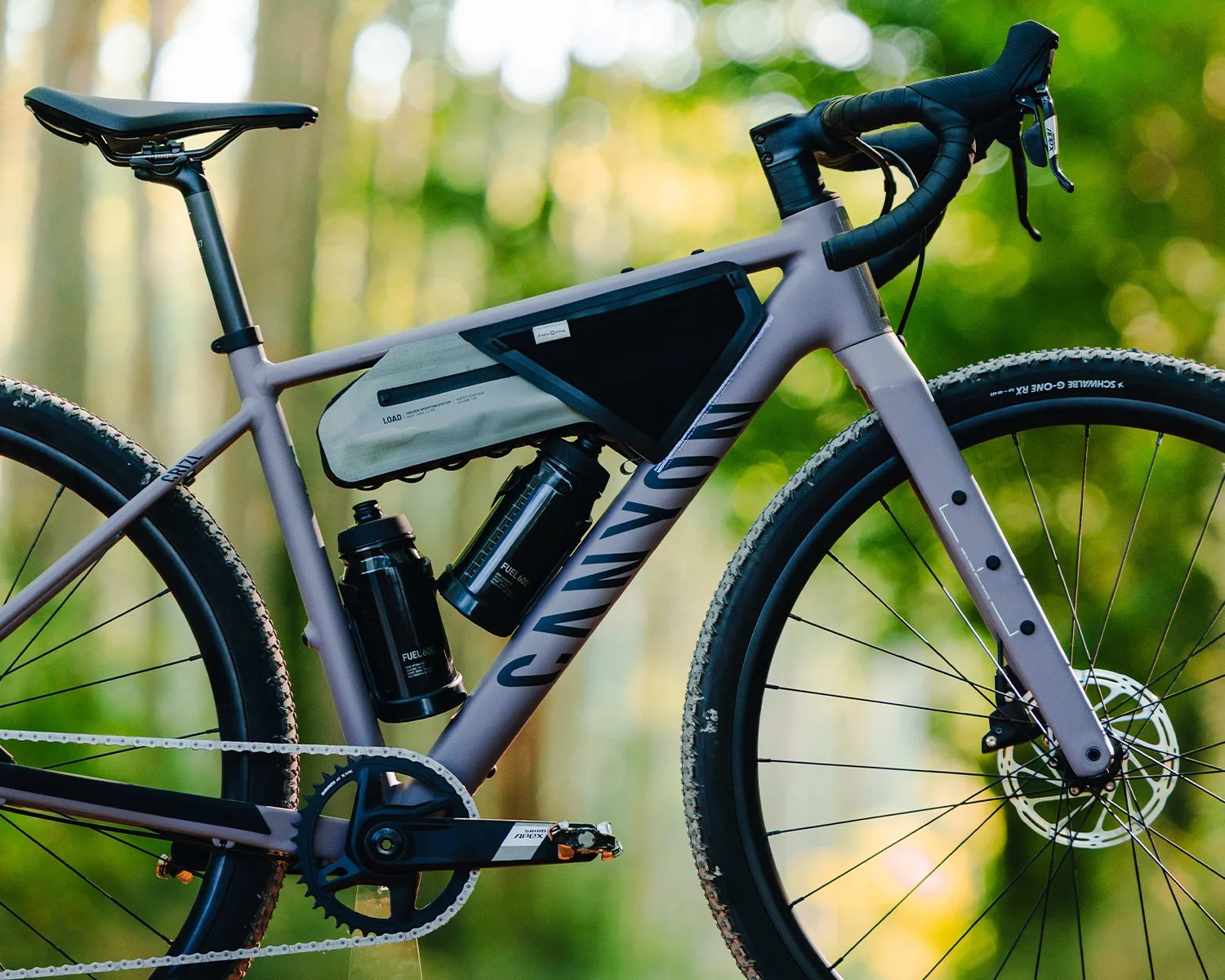 Matte gray Canyon gravel bike with black water bottles, frame bag, and drop handlebars in a forest setting.