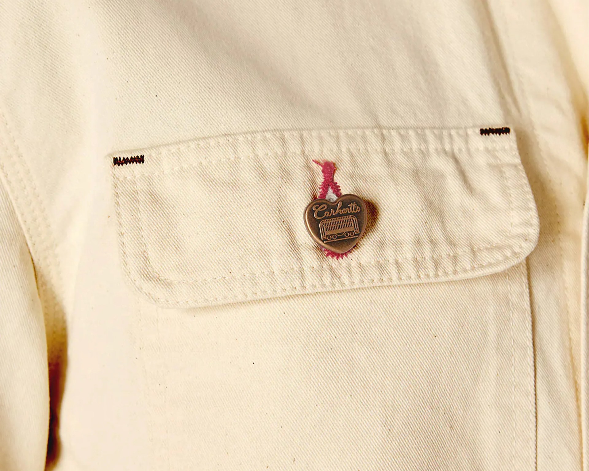 Close-up of a beige fabric pocket with a heart-shaped Carhartt button and red stitching.