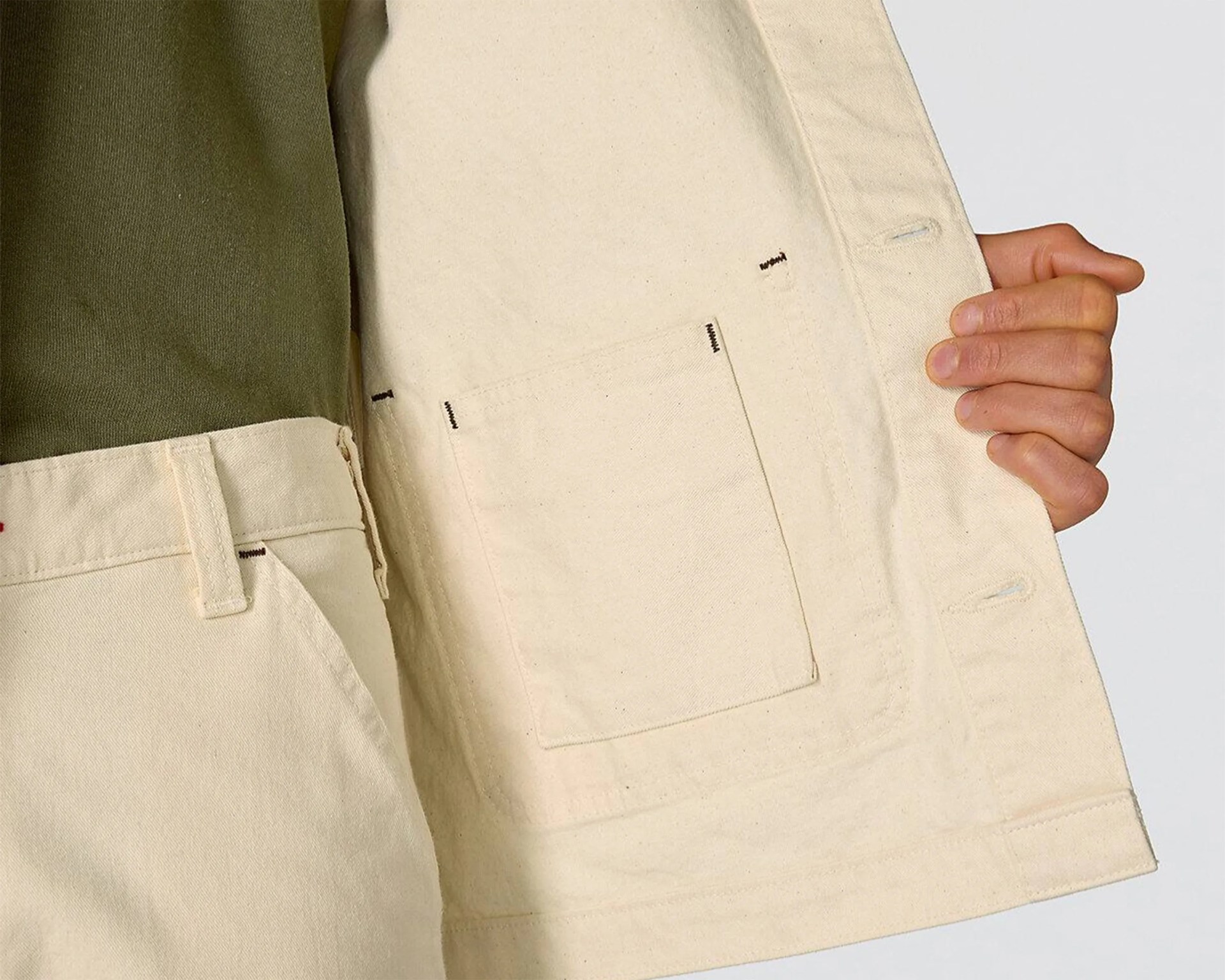 Close-up of a hand holding open the inside of a cream-colored jacket showing an inner pocket and matching cream pants with a belt loop.