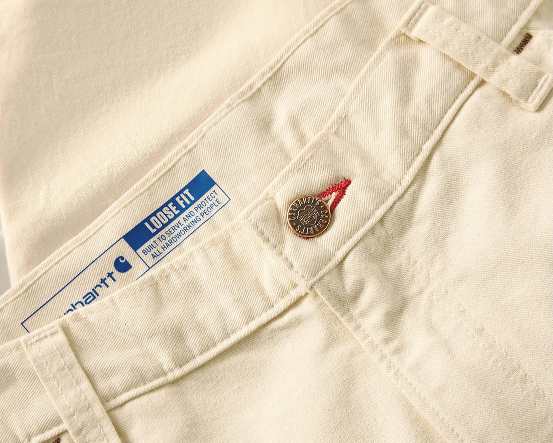 Close-up of cream-colored Carhartt pants with a metal button and blue label reading "LOOSE FIT.
