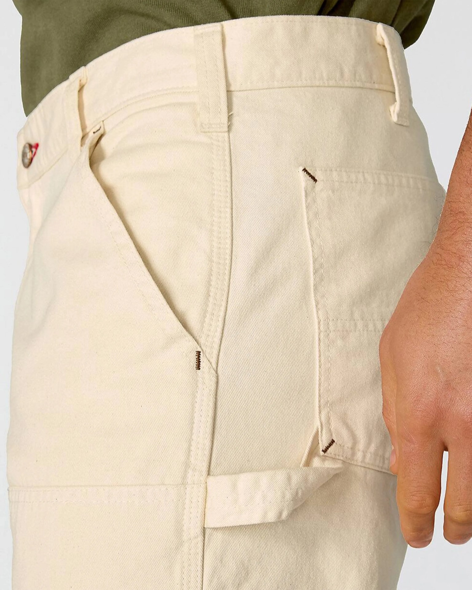 Close-up of beige pants with a side pocket and belt loops, worn with a green top and a hand visible.