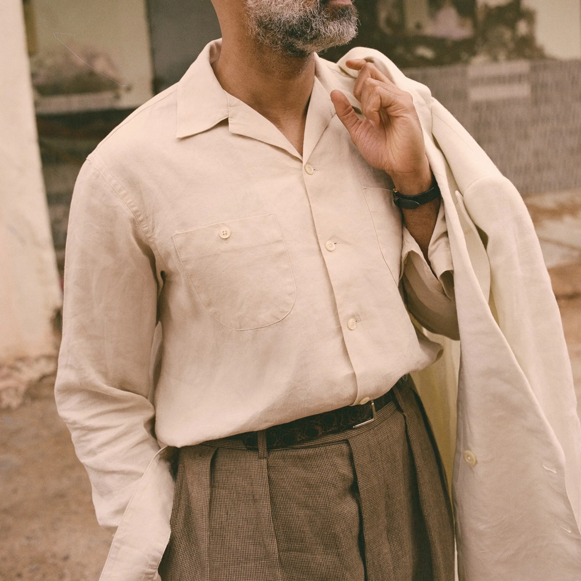 Beige button-up shirt with chest pockets worn tucked into brown plaid pleated trousers with a black belt.