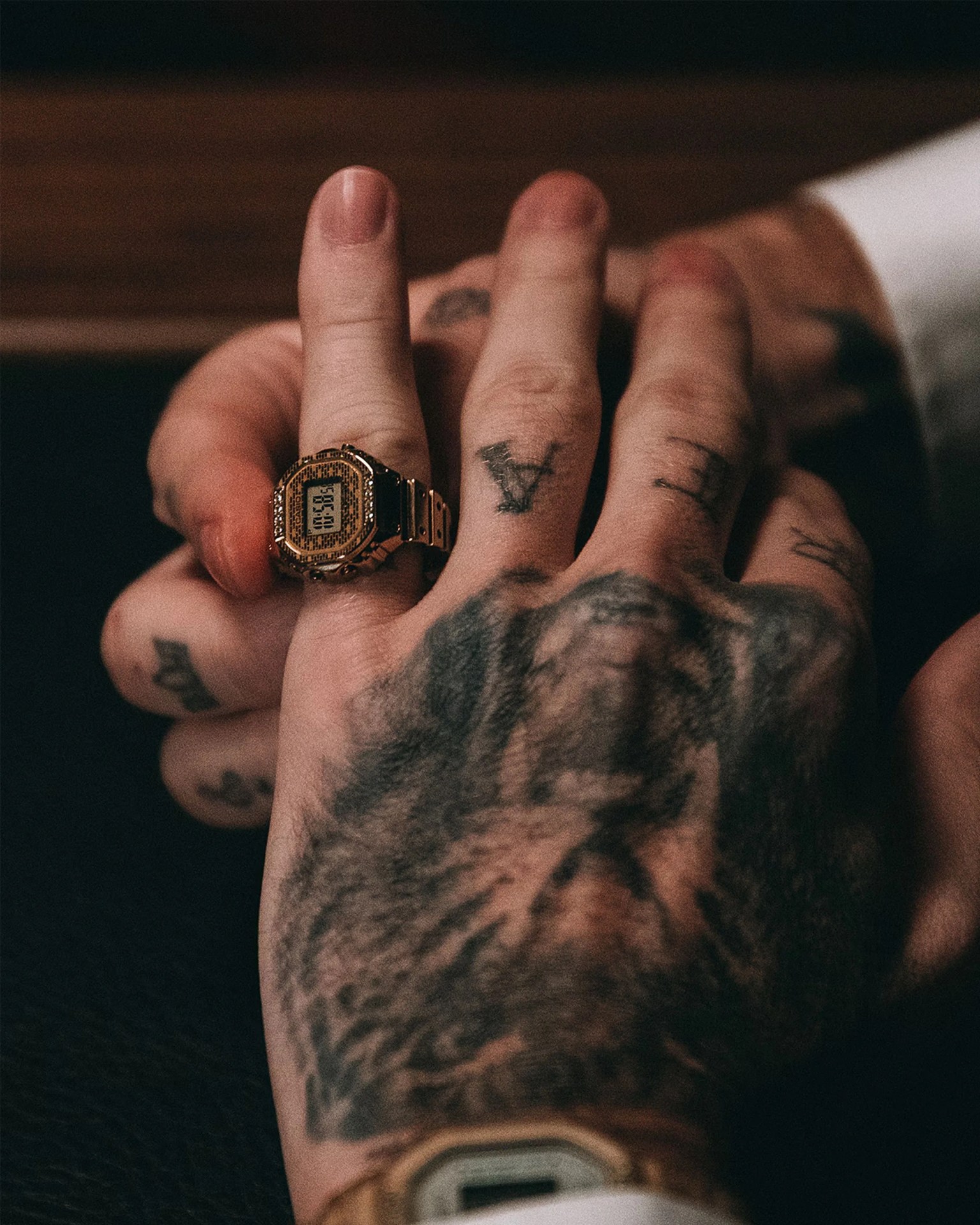 Hand with tattoos wearing a gold digital watch on the middle finger.