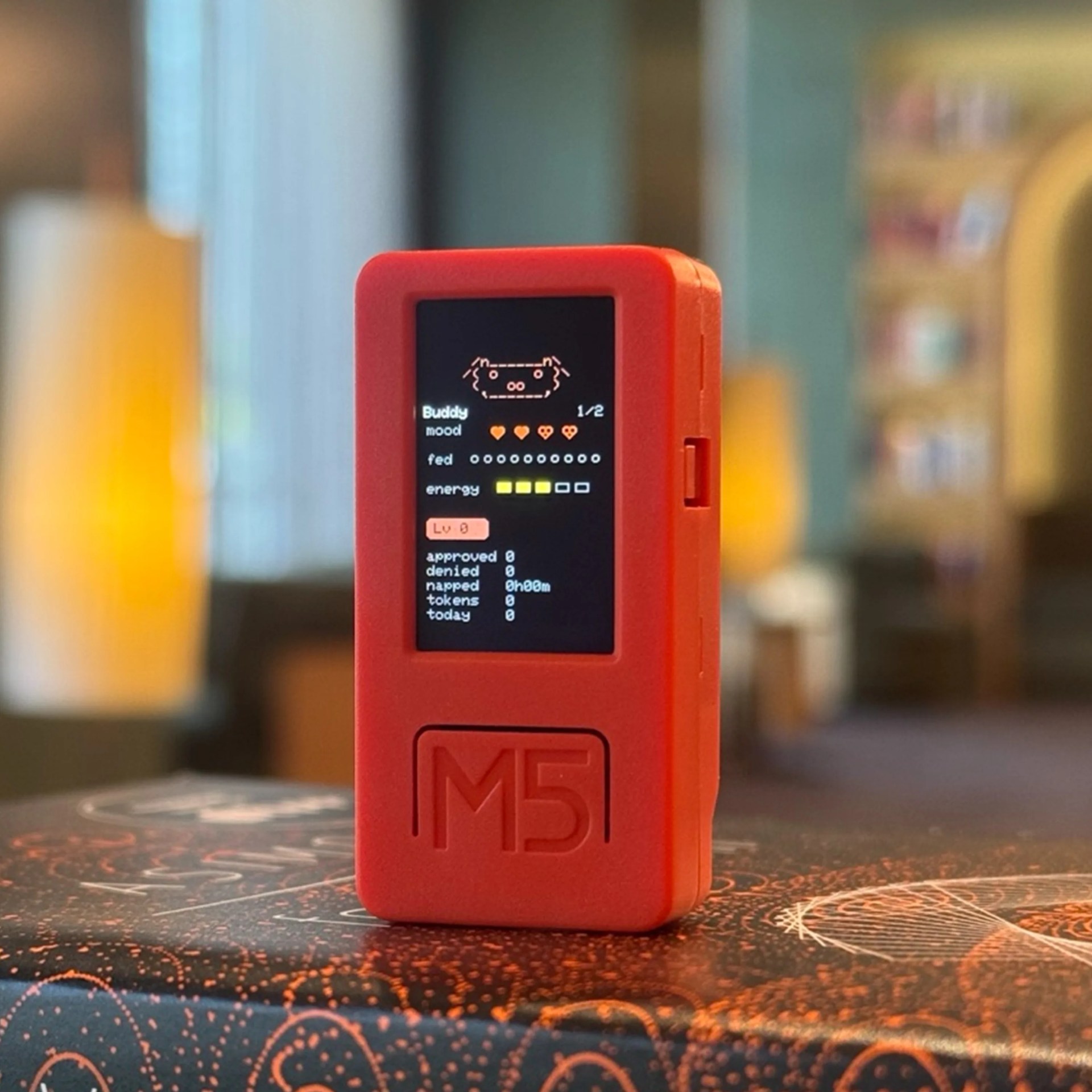 Red rectangular electronic device with a small screen displaying a pixel art character and status indicators, placed on a patterned surface.