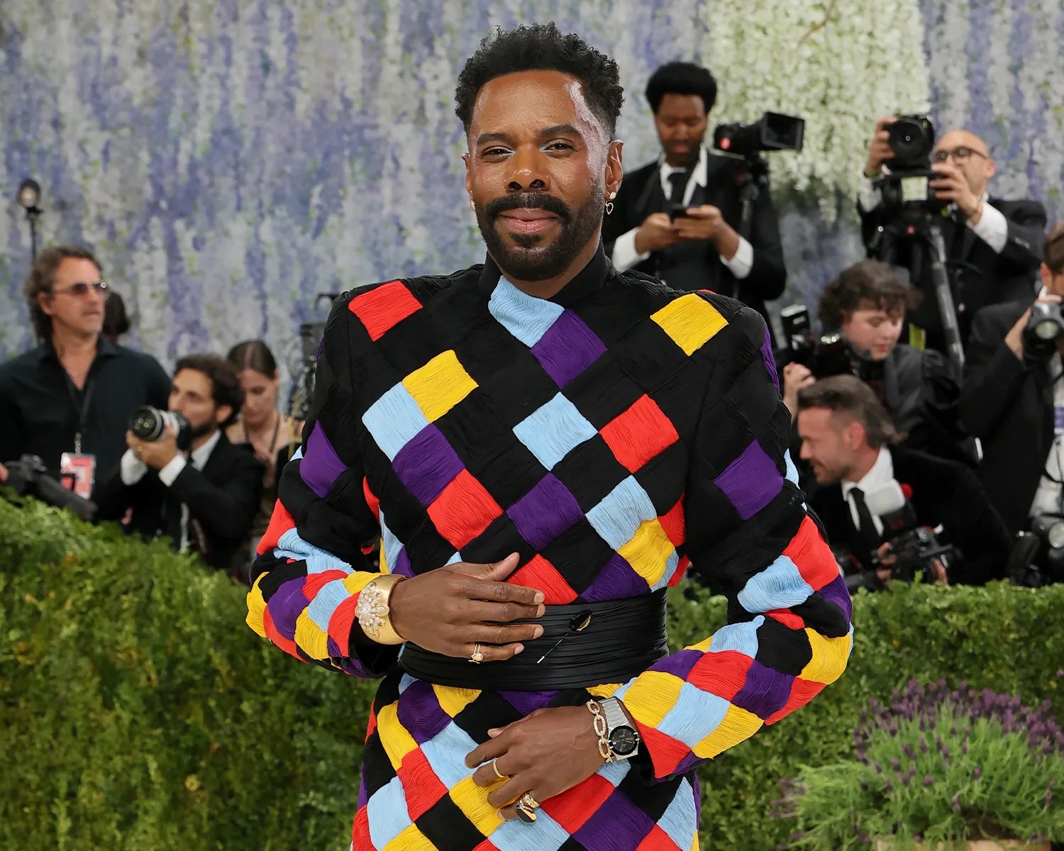Man wearing a multicolored checkered jacket with padded shoulders and gold jewelry at an event with photographers in the background.
