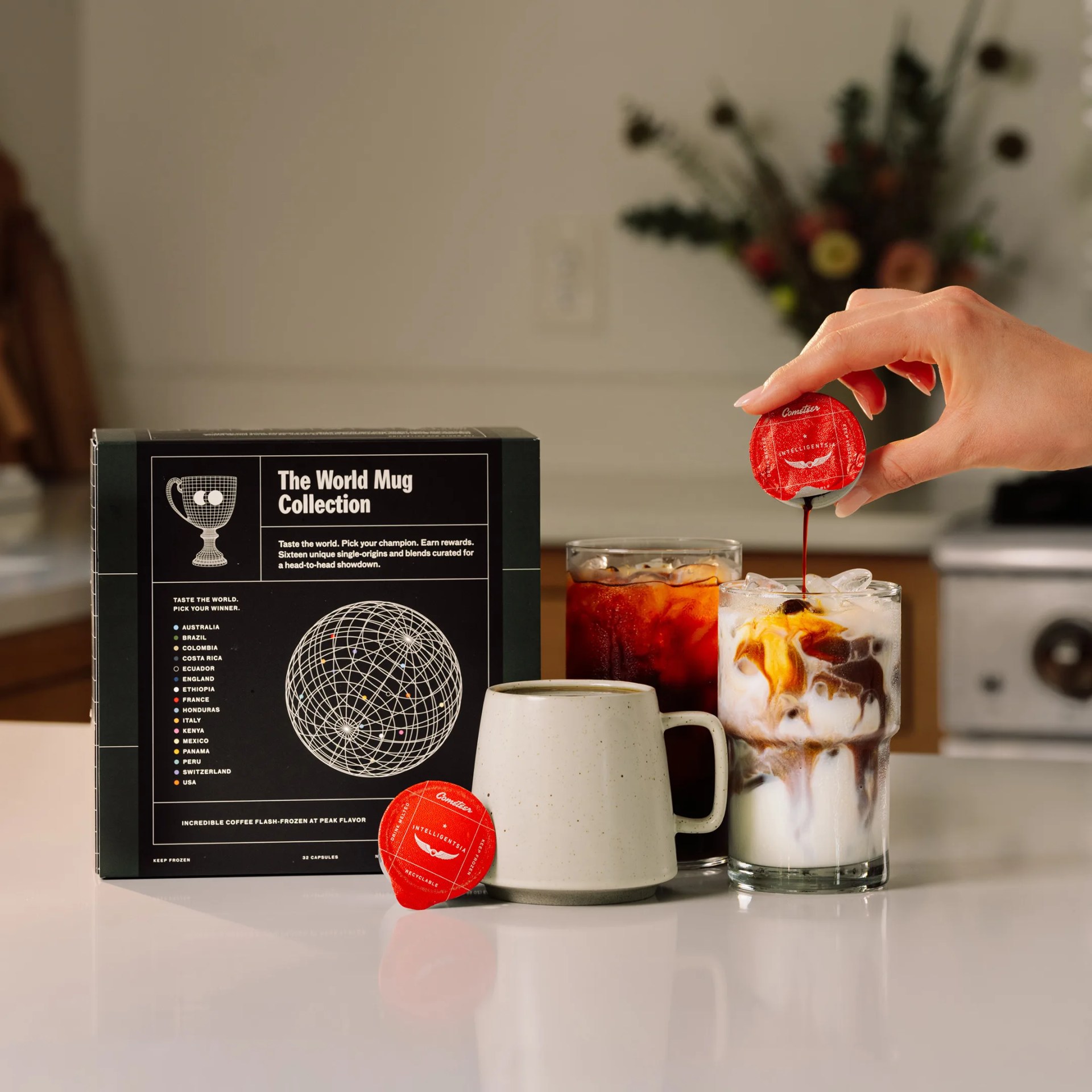Black box labeled "The World Mug Collection" with a white ceramic mug, two iced coffee glasses, and a hand pouring coffee from a red capsule.