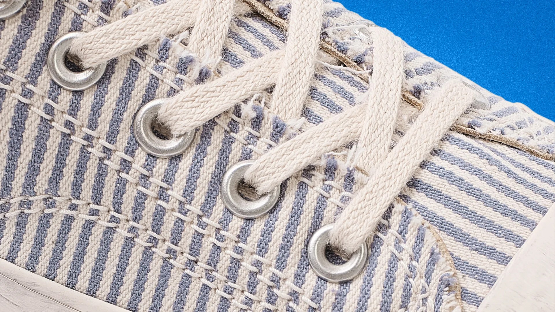 Close-up of beige and blue striped fabric sneaker with beige laces threaded through silver eyelets.