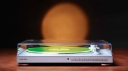 White Coolgeek turntable with a green vinyl record under a clear dust cover on a wooden surface.