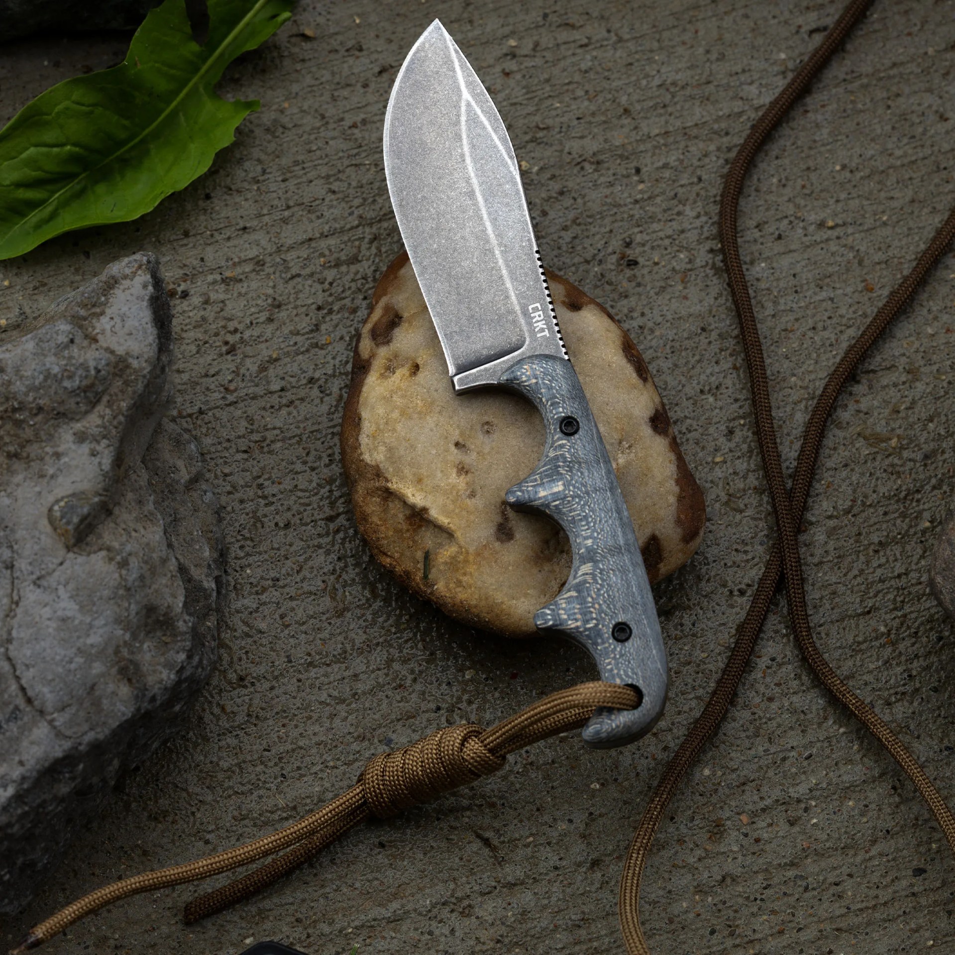Stonewashed fixed blade knife with gray textured handle and brown paracord lanyard on a rock and concrete surface.