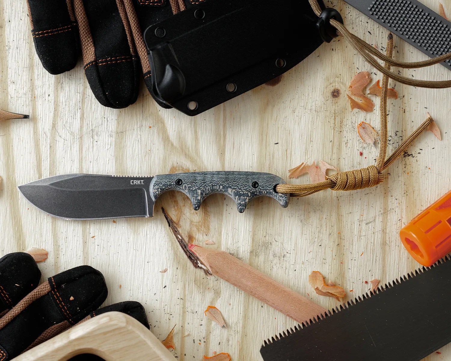Fixed-blade CRKT knife with textured gray handle and brown lanyard on a wooden surface with gloves and sawdust.