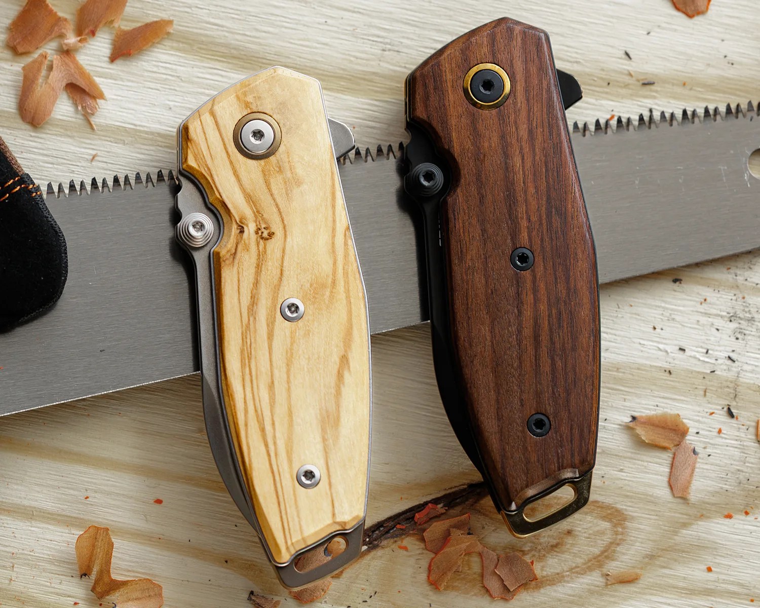 Two folding knives with wooden handles, one light and one dark, resting on a saw blade on a wooden surface.