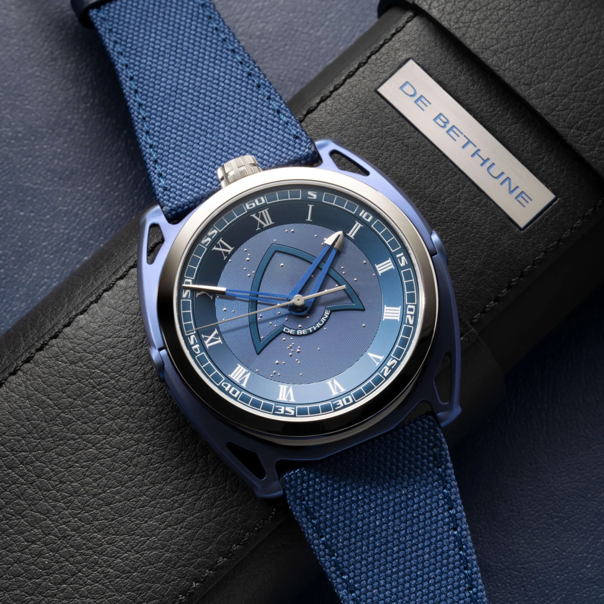 Blue fabric strap wristwatch with blue dial and silver bezel on black leather surface with "DE BETHUNE" label.
