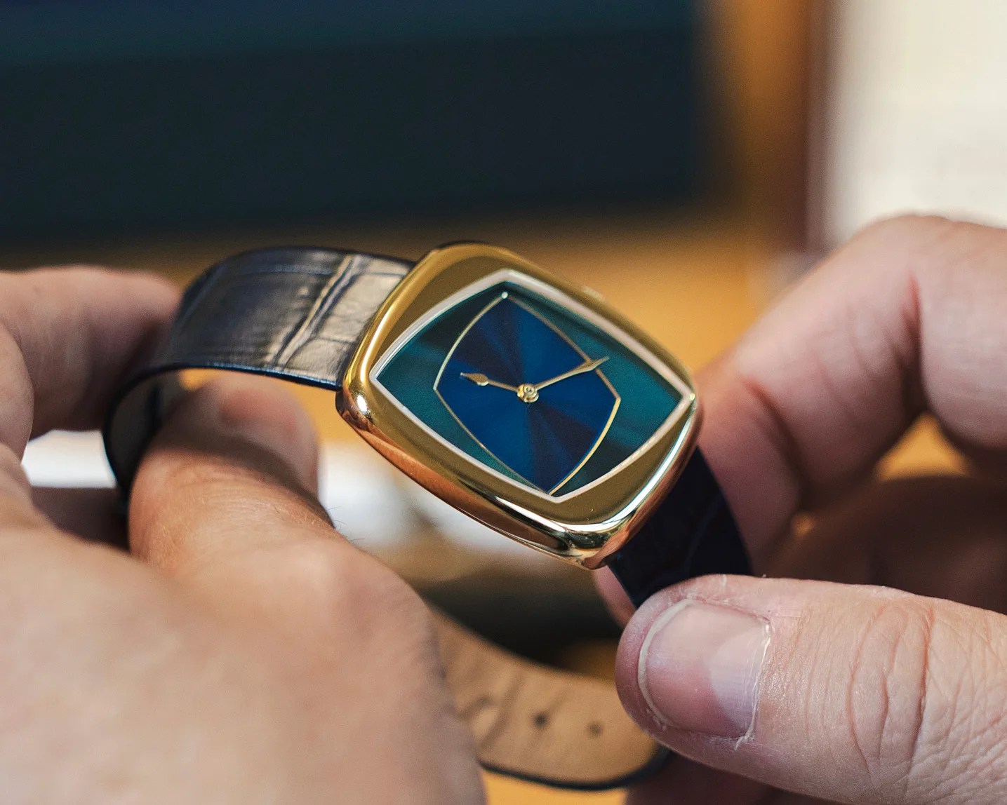 Gold rectangular wristwatch with a blue dial and black leather strap held between two hands.