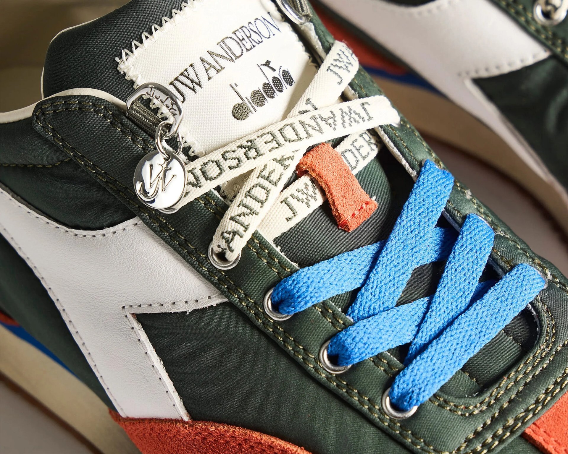 Close-up of a sneaker with green fabric, white leather accents, orange suede toe, blue laces, and J.W. Anderson branded cream laces and tongue label.