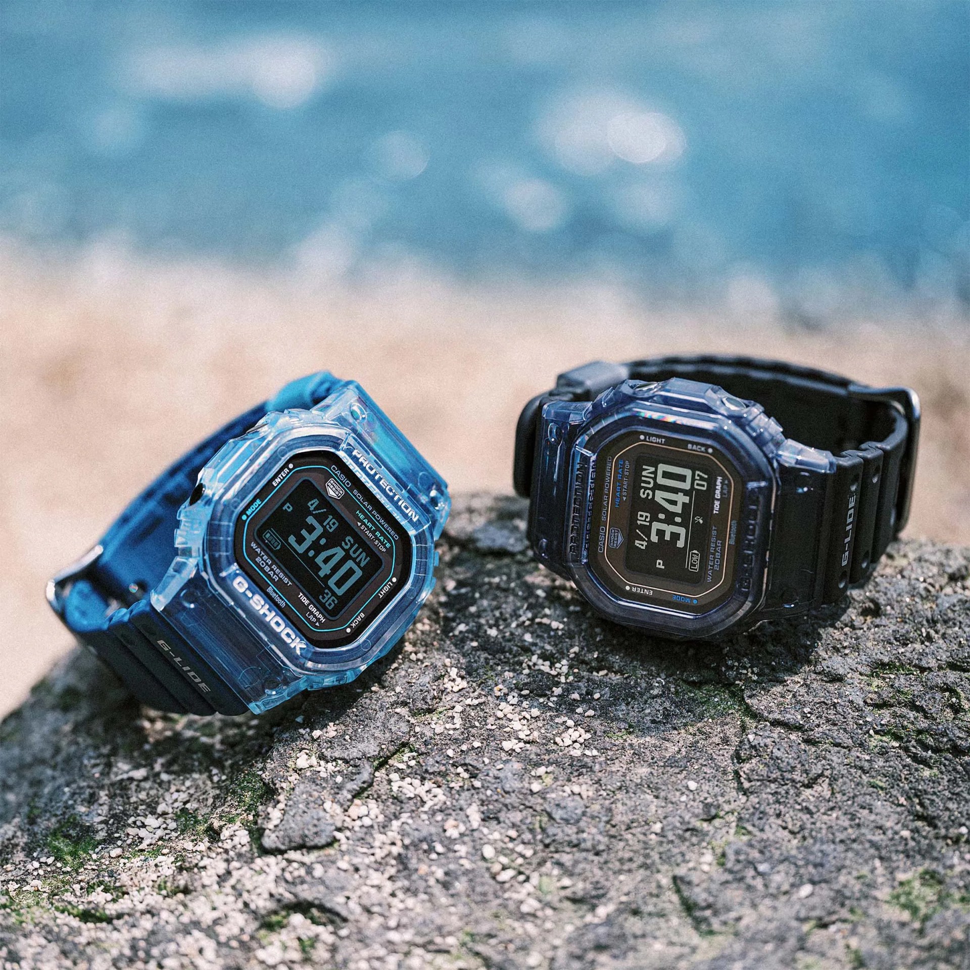 Two Casio G-Shock digital watches with translucent blue cases and black straps resting on a rocky surface near water.