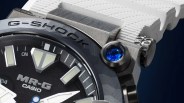 Close-up of a Casio G-Shock MR-G watch with a black dial, white hands, blue jewel on the case, and white textured strap.