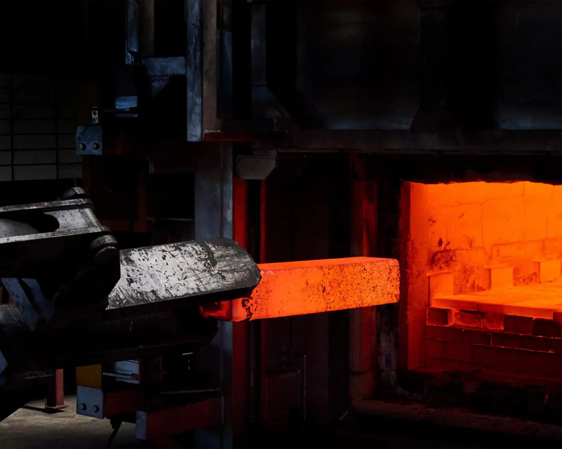 Glowing hot metal bar being processed in an industrial furnace with mechanical equipment.