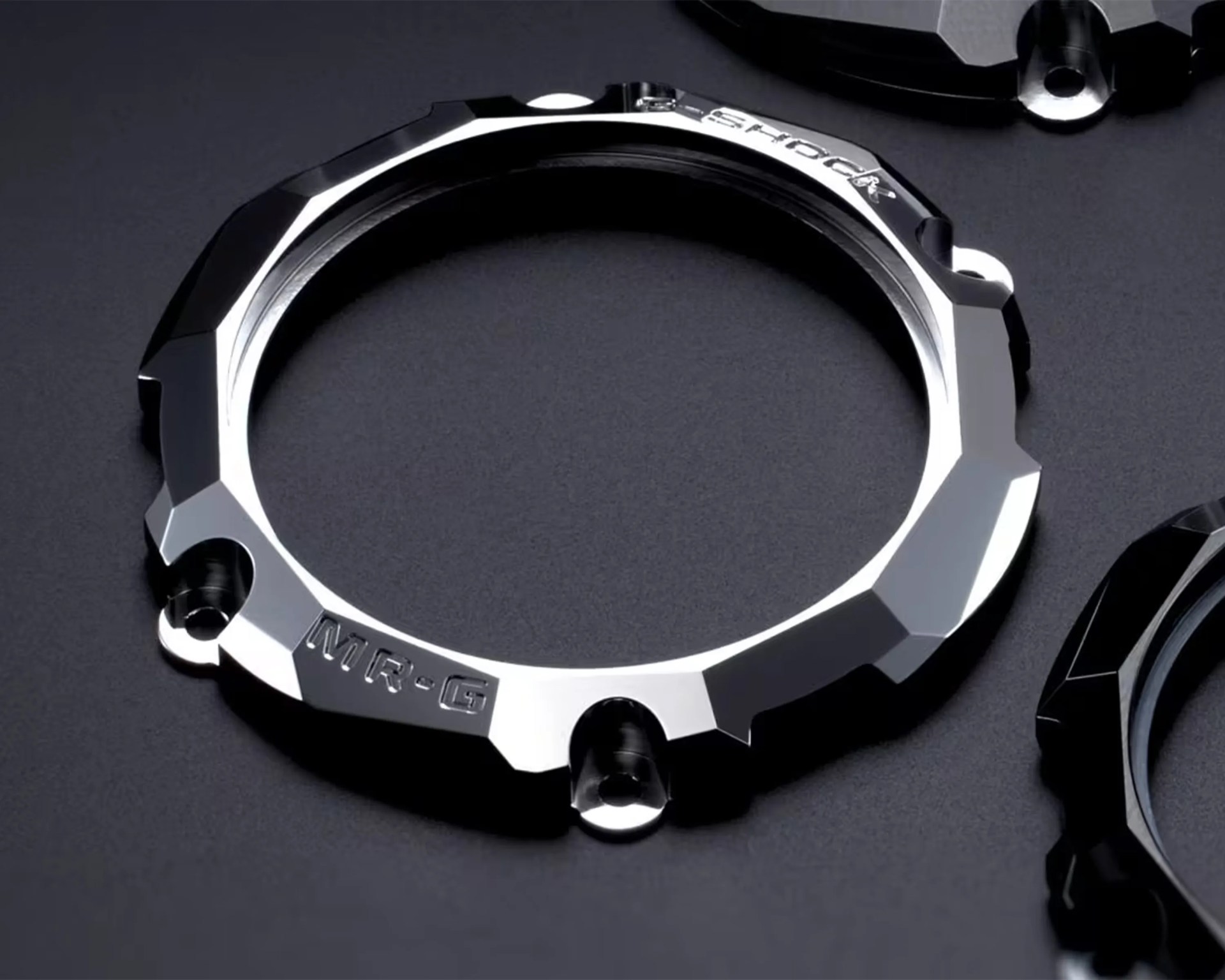 Polished silver metal watch bezel with "MR-G" and "SHOCK" engravings on a dark surface.