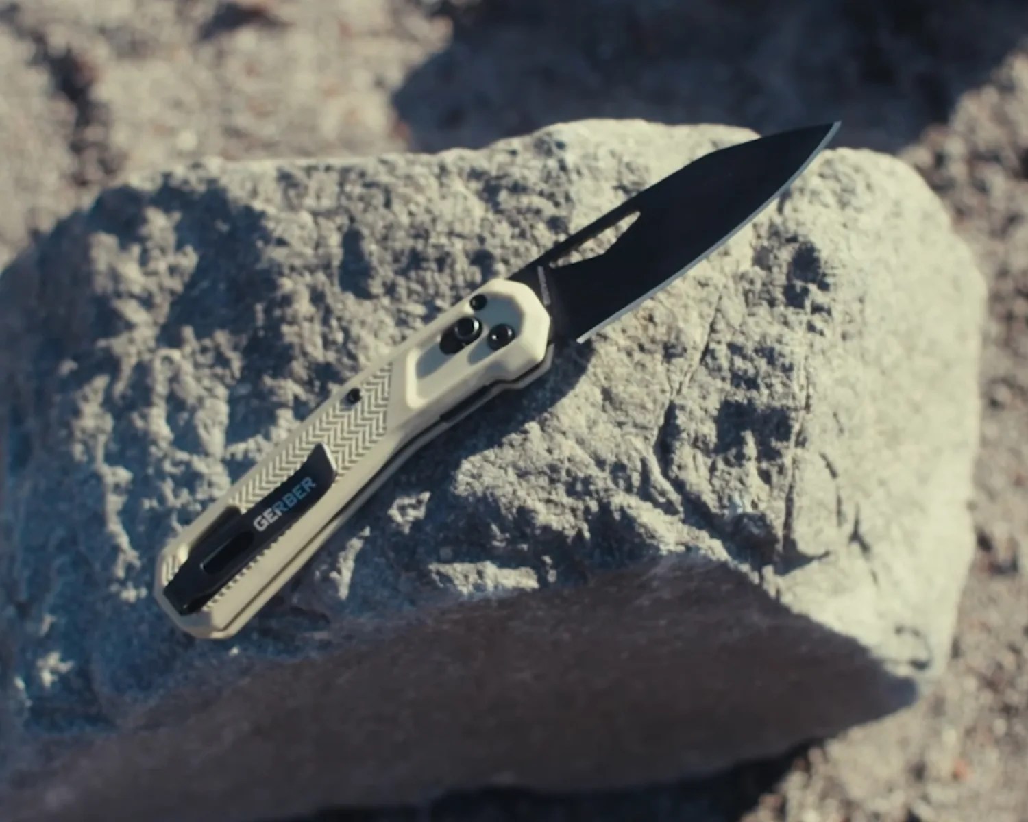 Folding knife with a black blade and tan textured handle labeled "Gerber" resting on a rock.