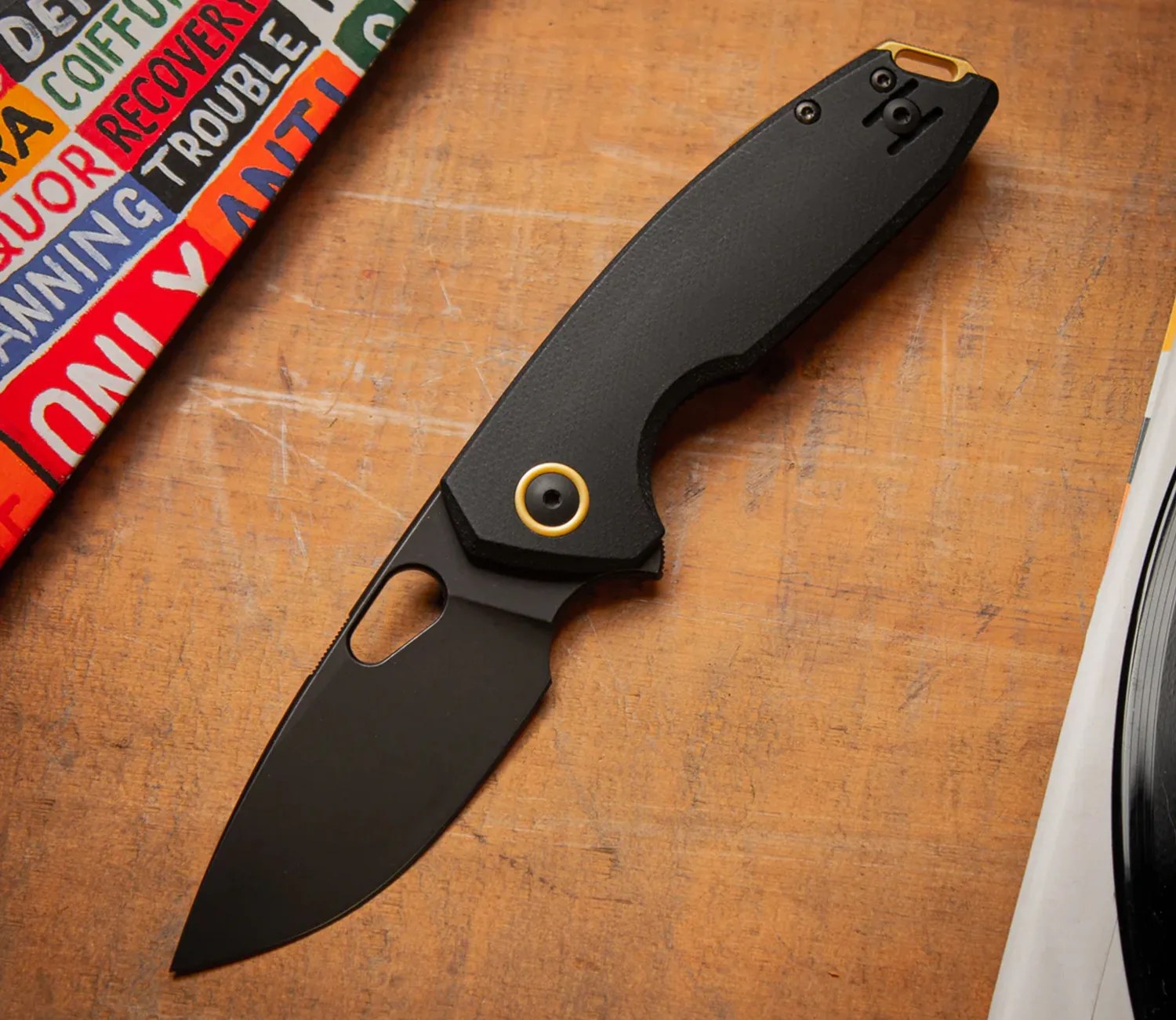 Black folding knife with textured handle and gold accent on a wooden surface next to colorful book and vinyl record.