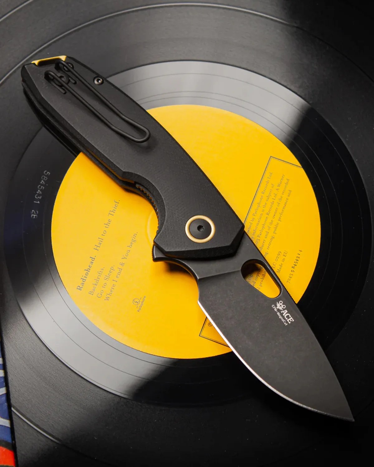 Black folding knife with a clip and partially serrated blade resting on a vinyl record with a yellow label.