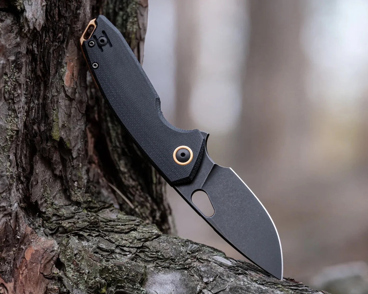 Black folding knife with a textured handle and gold accents stuck in tree bark.