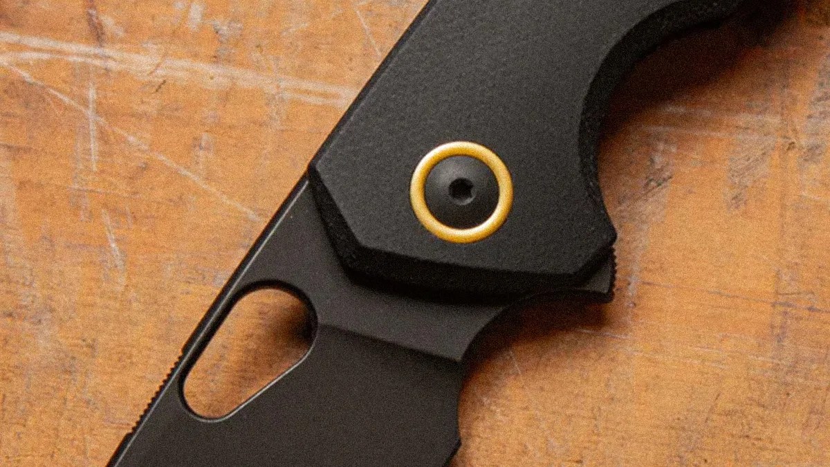 Close-up of a black folding knife handle with a gold circular pivot on a wooden surface.
