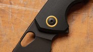 Close-up of a black folding knife handle with a gold circular pivot on a wooden surface.