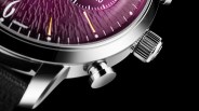 Close-up of a silver watch case with a textured purple dial, crown, and push button on a black background.
