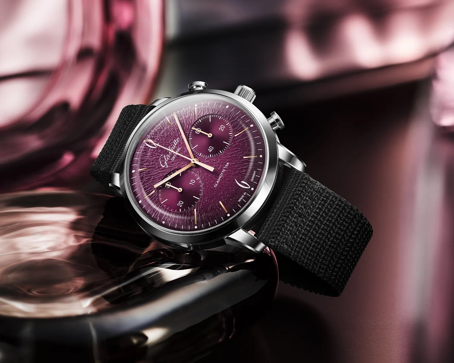 Silver chronograph wristwatch with textured purple dial and black woven strap on reflective surface.