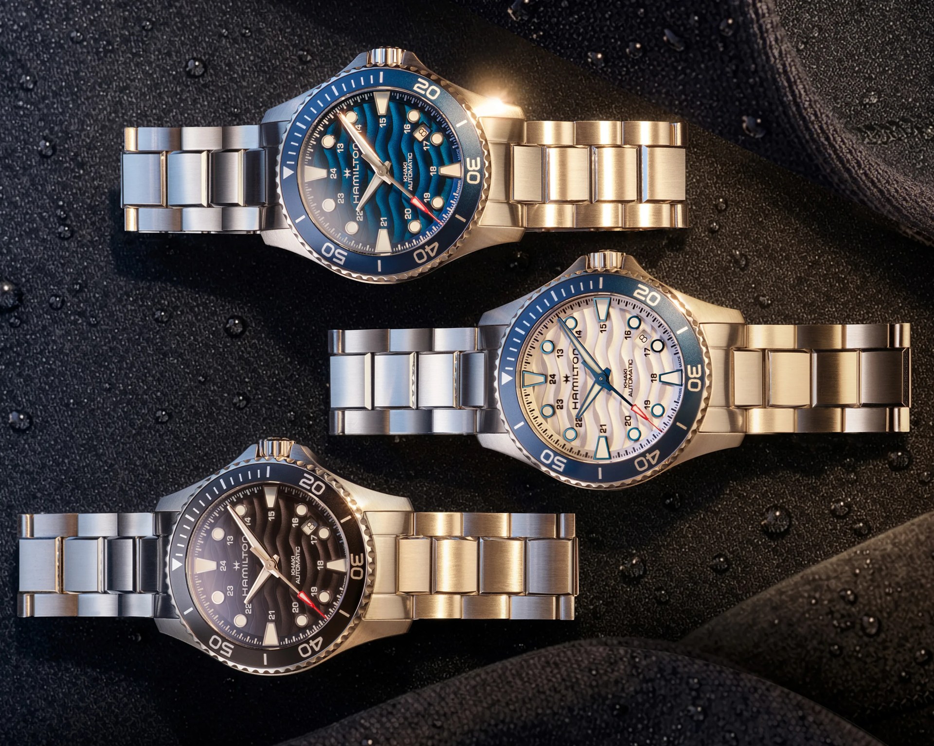 Three Hamilton Khaki Automatic watches with stainless steel bracelets in blue, white, and black dials on a wet black surface.