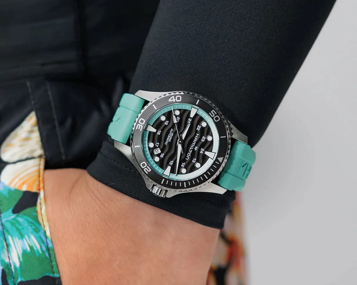 Hamilton wristwatch with a black dial, wave pattern, black bezel, and turquoise rubber strap worn on a wrist.