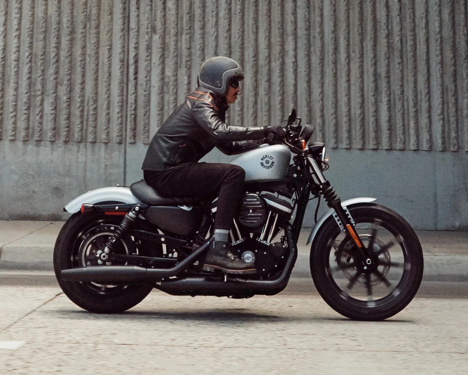 Person wearing a black helmet and leather jacket riding a silver Harley-Davidson motorcycle on a street.