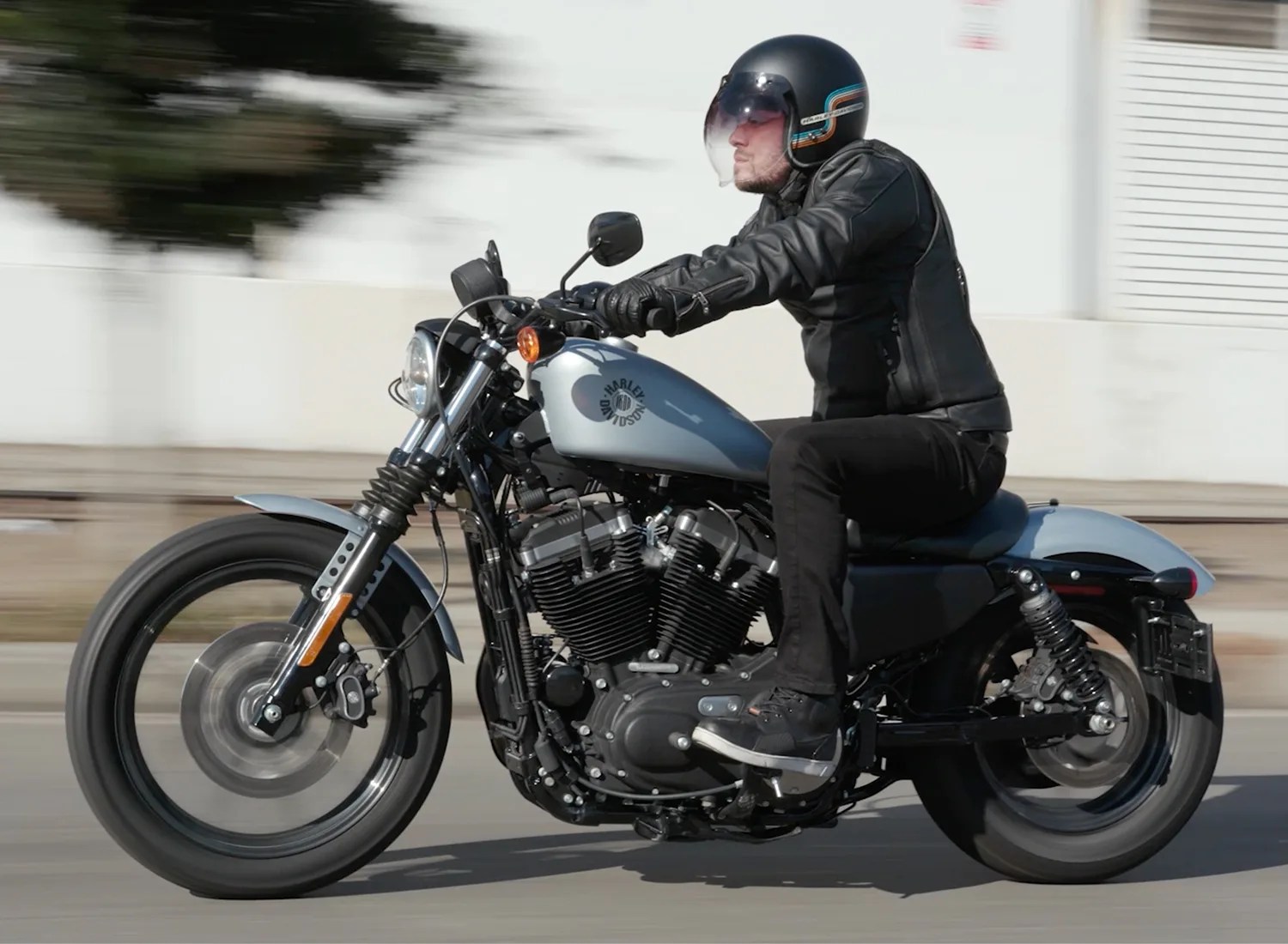 Person wearing black leather jacket and helmet riding a silver Harley-Davidson motorcycle.