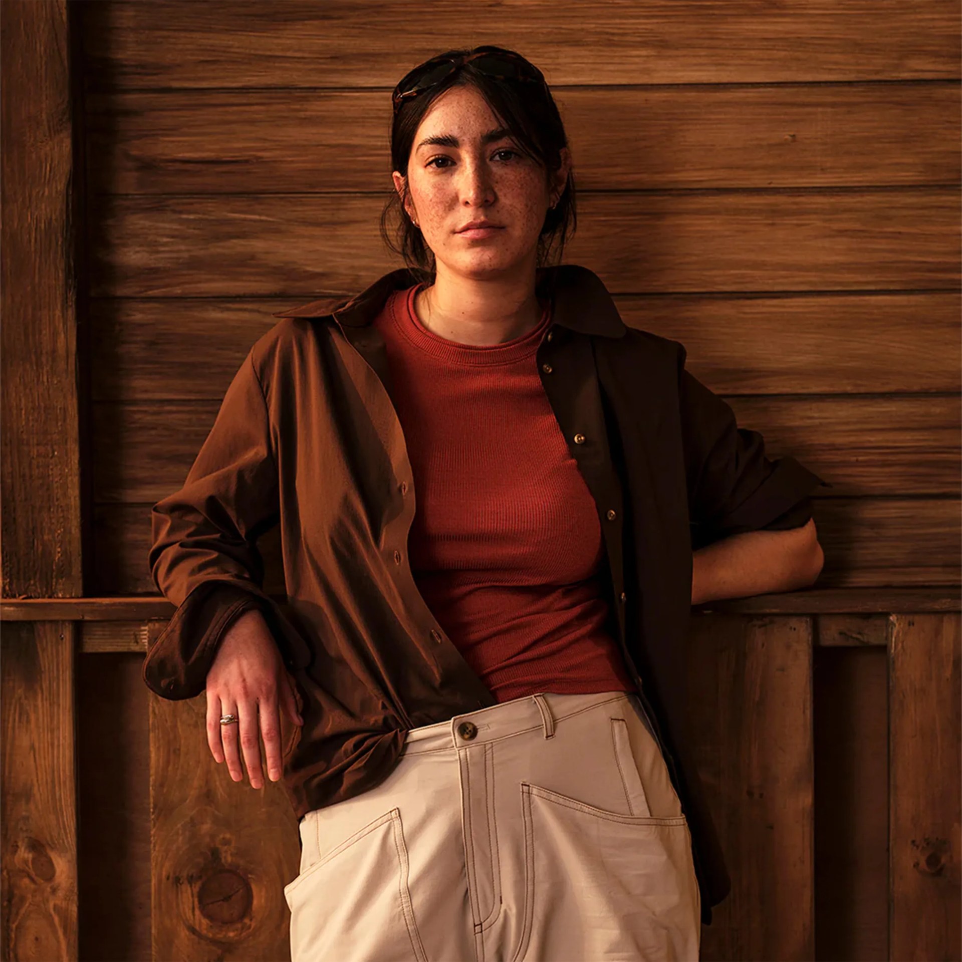 Woman wearing a brown button-up shirt over a rust-colored top and beige pants, standing against a wooden wall.
