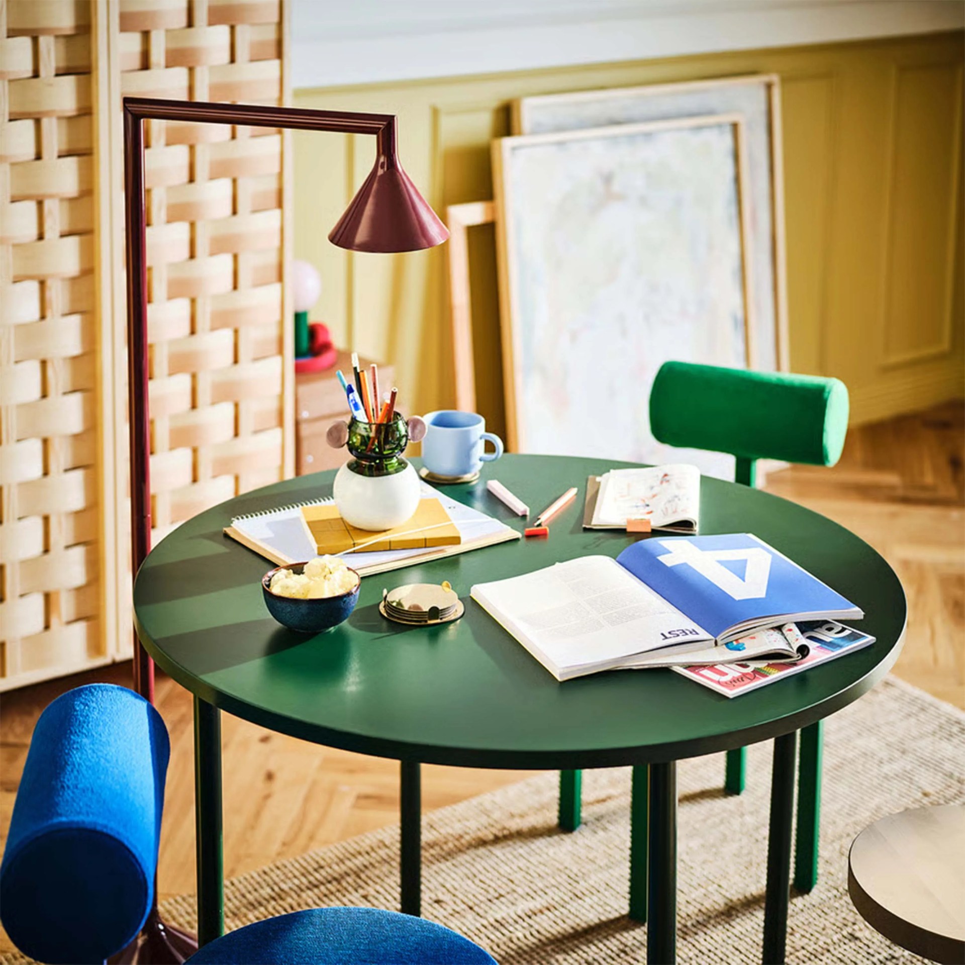 Green round table with books, pencils in a vase, a blue mug, and a red desk lamp with a green and blue chair.
