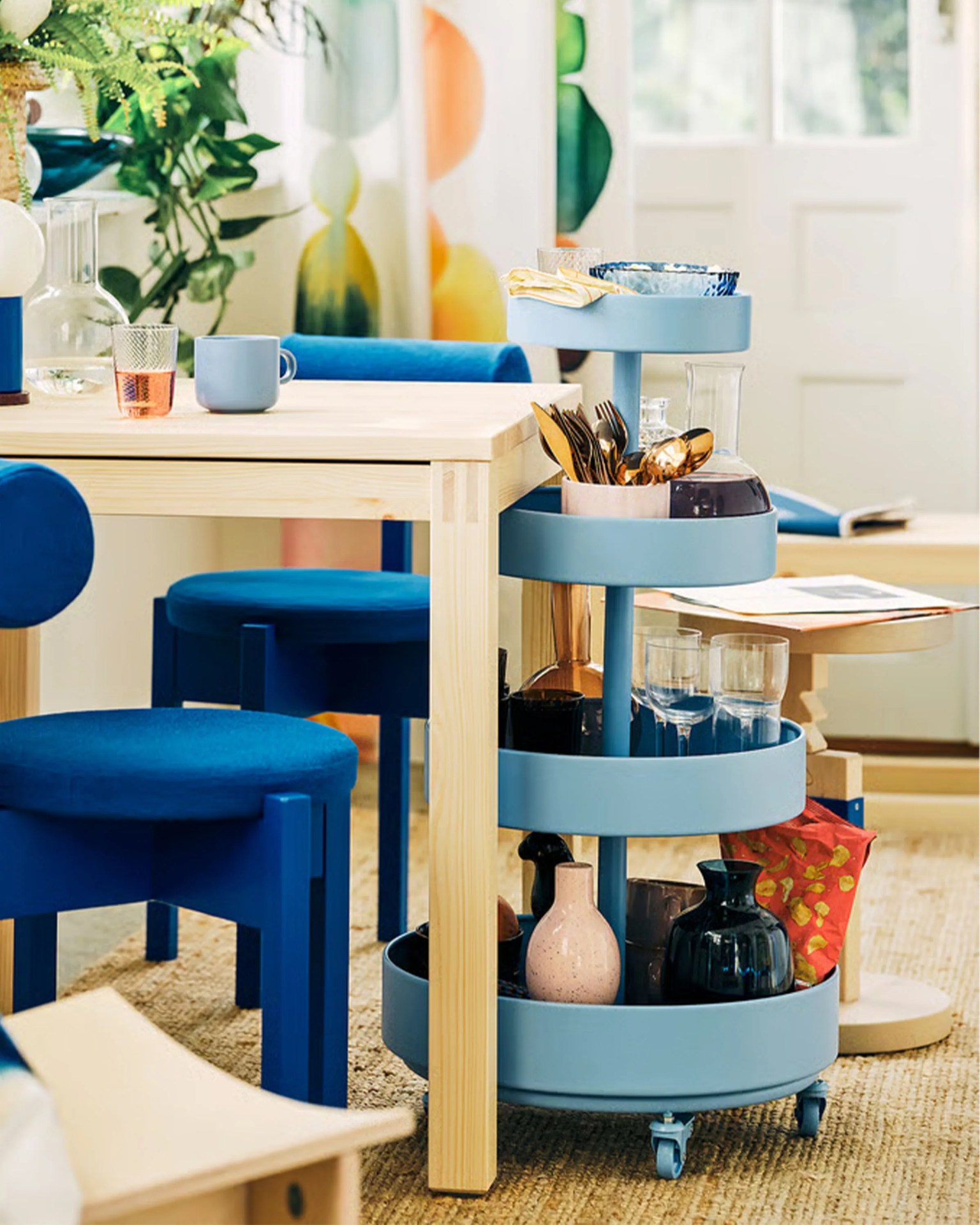 Light blue three-tier rolling cart holding glassware, utensils, and bottles next to a light wood dining table with blue velvet chairs.