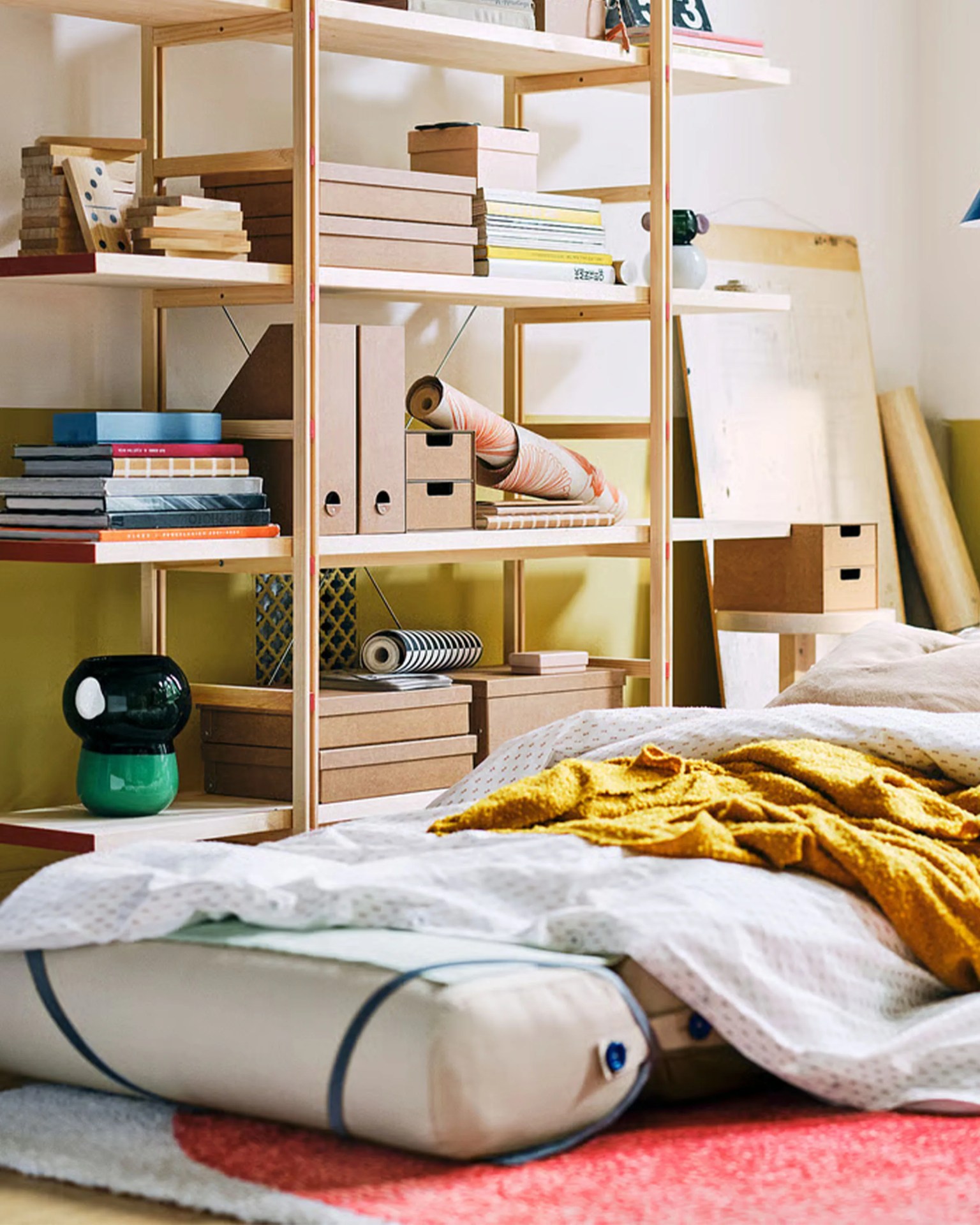 Wooden shelving unit with books, cardboard boxes, rolled papers, and a green and black ceramic vase next to a bed with white bedding and a mustard yellow blanket.