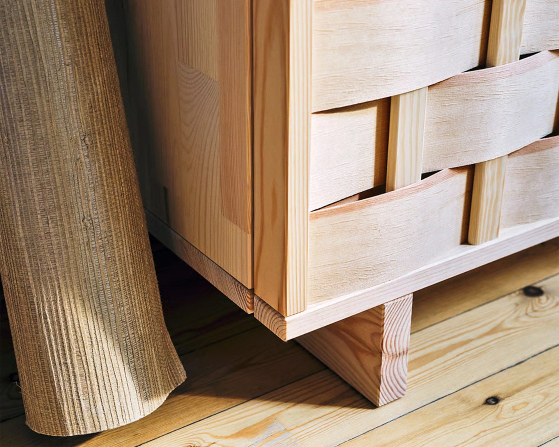 Close-up of light wood furniture corner with woven wooden slats and textured beige fabric on wooden floor.