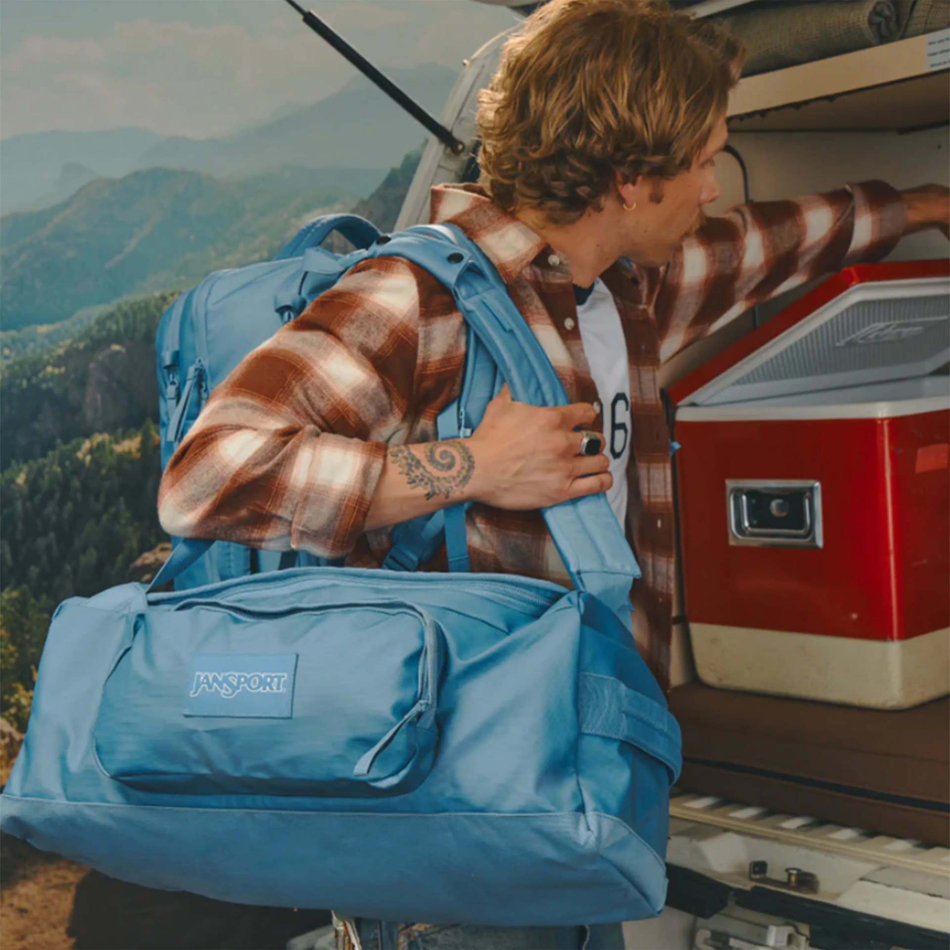 Light blue Jansport duffel bag worn by a person with a blue backpack and plaid shirt near a red cooler.